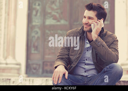 Man talking on a phone. Casual professional entrepreneur using smartphone smiling outside old building. Outdoor portrait of modern guy with mobile in  Stock Photo