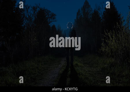 Male silhouette at the dark forest pathway through the bushes in the night. Man standing on the road against the car headlights. Mystery ghost concept Stock Photo