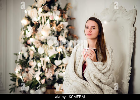 Young beautiful woman with sitting home holding cup of hot coffee wearing knitted warm sweater. Christmas tree decorations and lights in the room Stock Photo