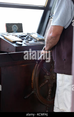 a man or the captain of a ship or boat holding a ship's wheel or helm whilst driving a ship or steering a boat. nautical, maritime ships bridge. Stock Photo