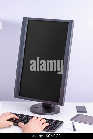 Black blank pc monitor turned vertical and hand on keyboard on desk. Stock Photo