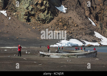 Adventure tourists walk near long abandoned water boats from the whaling era on a volcanic beach at Whalers Bay, Deception Island, South Shetland Islands Stock Photo