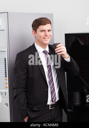 Portrait of happy businessman drinking coffee standing near vending machine Stock Photo