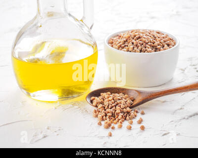 Brown buckwheat in spoon and buckwheat oil in glass bottle on white wooden background. Copy space. Stock Photo