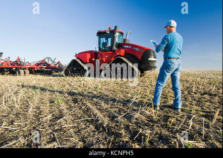 farmer using a tablet in front of a tractor and air seeder planting winter wheat in a zero till field containing canola stubble, near Lorette, Manitoba, Canada