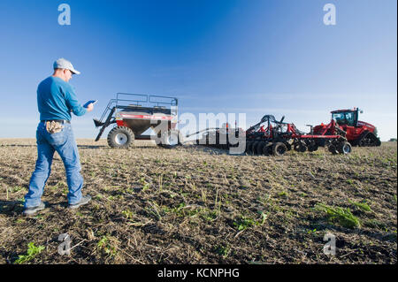 farmer using a tablet in front of a tractor and air seeder planting winter wheat in a zero till field containing canola stubble, near Lorette, Manitoba, Canada