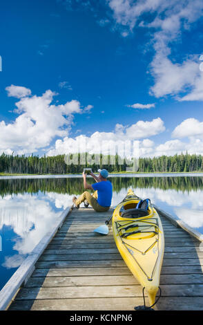 man sitting on dock next to his kayak while using a tablet, Two Mile Lake , Duck Mountain Provincial Park, Manitoba, Canada Stock Photo