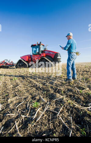 farmer using a tablet in front of a tractor and air seeder, planting winter wheat in a zero till field containing canola stubble, near Lorette, Manitoba, Canada