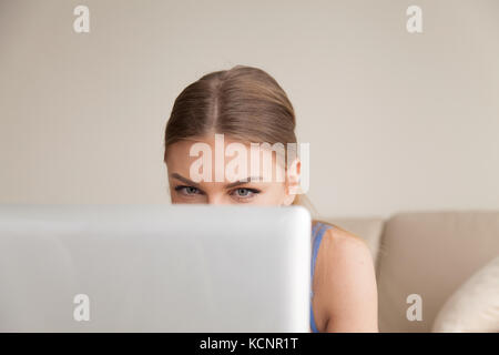 Curious teen using computer, young funny girl peeking over top of laptop, looking at screen, playing interesting online game, having fun while chattin Stock Photo