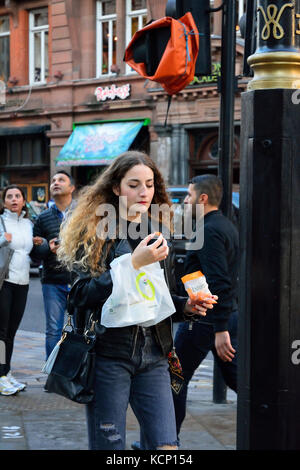young woman wearing black leather biker jacket eating a snack whilst walking in the street in London, England, UK Stock Photo