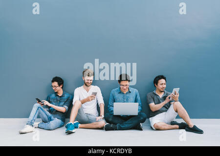 Multiethnic group of four men using smartphone, laptop computer, digital tablet together with copy space on blue wall. Lifestyle with gadgets concept Stock Photo
