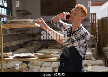 Male potter taking on mobile phone while working in pottery workshop Stock Photo
