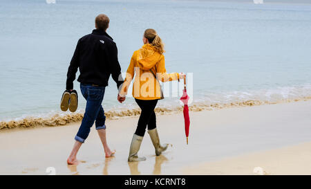 Coupler walking on Porthminster beach holding hands. Man walking barefoot while woman weras wellington boots and carries umbrella. Stock Photo