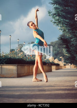 Dancing ballerina in blue ballet tutu and point on european street with trees in summer or autumn. Young beautiful brunette woman practicing stretchin Stock Photo