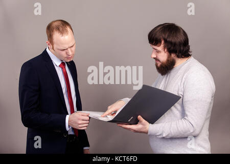 An employee in a gray jumper shows the boss in a suit his plan for the development of the company in a gray folder, two men look in a folder on a gray Stock Photo