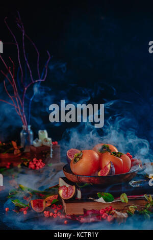 Autumn harvest still life with persimmons and figs on a stack of magical books. Dark Halloween concept with smoke. Dark food photography with copy spa Stock Photo