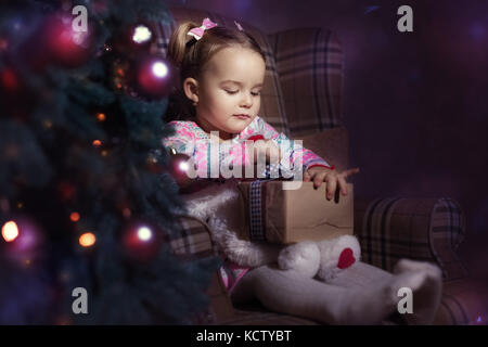 Baby girl sitting in a chair near a Christmas tree. Girl holding a gift, she tries to open it Stock Photo