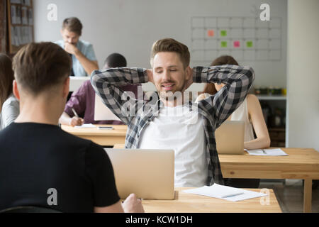 Smiling young male entrepreneur relaxing stretching back at workplace in shared office. Taking a break from heavy workflow, chilling after positive me Stock Photo