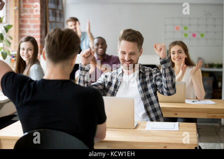 Young casually dressed male office worker throws hands in air celebrating achievement at work. Coworkers around cheer and clap hands. Rewarding outcom Stock Photo