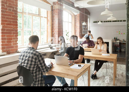Young casually dressed employees at work in coworking office. Corporate team working on laptops and taking notes in shared room, brainstorming about n Stock Photo