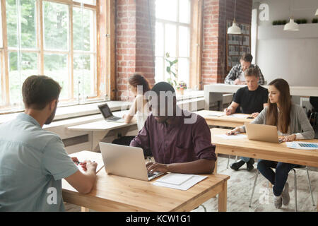 Multiethnic group of casual coworkers at work in shared loft office. Corporate team working on laptops, taking notes in shared room, brainstorming abo Stock Photo