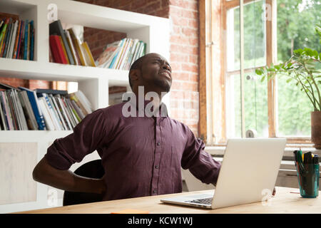 Young exhausted african american male office worker having back discomfort at work desk in office. Fatigued casual businessman feeling back pain after Stock Photo