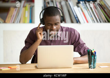 Smiling african american office worker in headphones looking at laptop screen. Young  casual businessman studying foreign language, communicating with Stock Photo