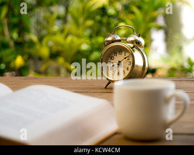 text book with vintage alarm clock and White coffee cup on the wood table. The background is green from tree and light bokeh Stock Photo