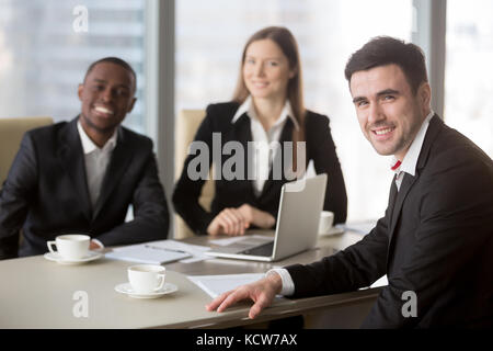 Portrait of friendly caucasian businessman sitting at negotiation table with multinational colleagues or partners during meeting. Interracial team of Stock Photo