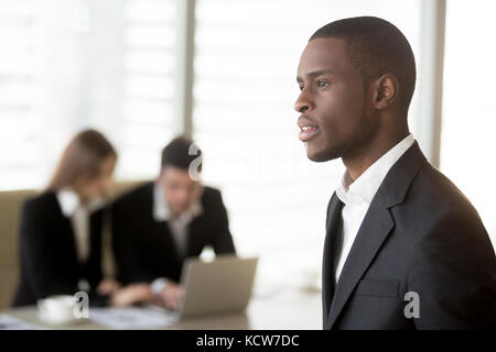 Side view portrait of handsome black businessman looking in distance, caucasian businesspersons working on laptop at office desk on background. Employ Stock Photo