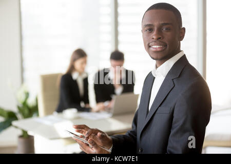 Portrait of smiling african american businessman using digital tablet and looking at camera, caucasian colleagues using laptop on background. IT start Stock Photo