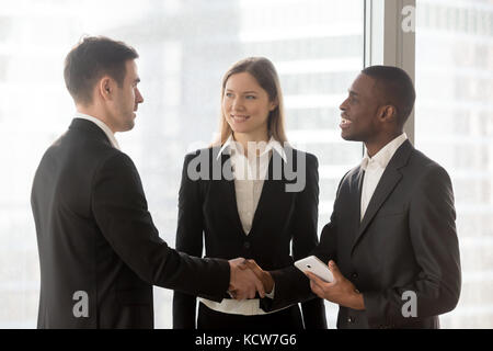 African american and caucasian business partners shaking hands after successful negotiation in office. Young female secretary standing between two com Stock Photo