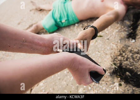 Woman puts a finger of a man to unlock his phone. Man is sleeping on the sand. A woman is stealing a phone. Stock Photo