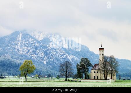 Old church with high bell tower and graveyard bellow mountains.  April weather. Wet spring snow in already fresh green grass  of meadow bellow mountai Stock Photo