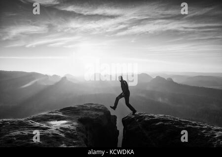Crazy hiker in black is jumping between rocky peaks. Happy man. Wonderful daybreak in rocky mountains, heavy orange mist in deep valley. Miracle of na Stock Photo