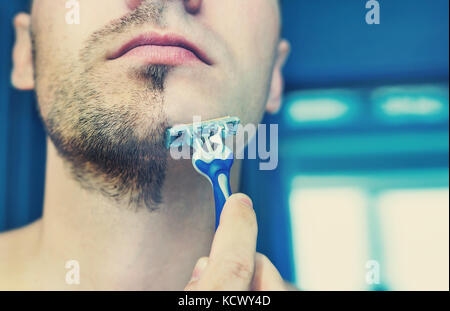 Young man shaves his short beard shaving machine tool. Half face with a beard half shaved. Stock Photo