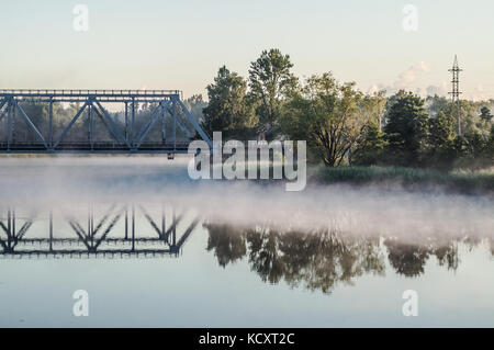 Railway bridge above misty lake. Reflection on water surface. Stock Photo