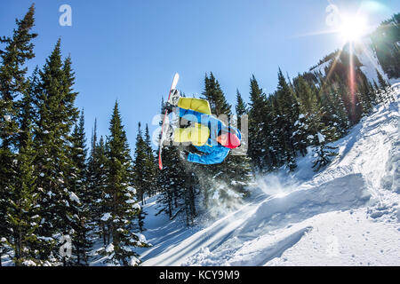 Snowboarder freerider jumping from a snow ramp in the sun on a background of forest and mountains. Stock Photo