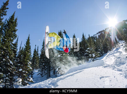 Snowboarder freerider jumping from a snow ramp in the sun on a background of forest and mountains.