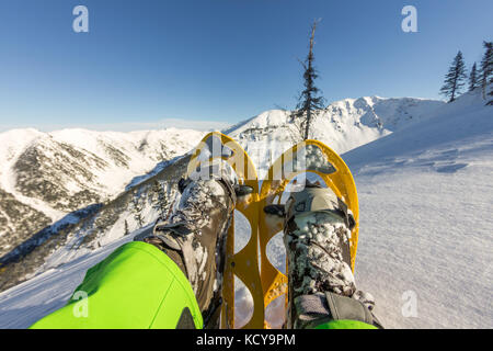 Winter sport activity concept. Hiker looking over his snowshoes at the beautiful landscape in the French mountains during sunset. Stock Photo