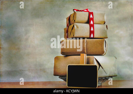 Gift boxes wrapped in rustic paper and heart shapes ribbon, small blackboard with place for text on abstract  background. Stock Photo