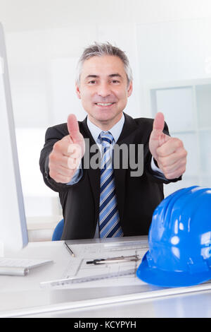 Happy Male Senior Architect Showing Thumbs Up Sign At Desk Stock Photo