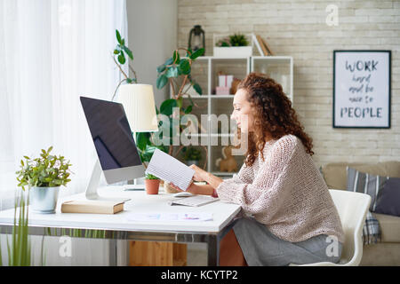 Side view portrait of curly haired mixed-race woman sitting at desk in modern apartment reading book Stock Photo