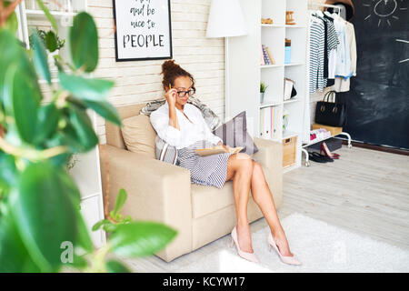 Portrait of stylish young woman wearing glasses reading book at home sitting in comfortable armchair Stock Photo