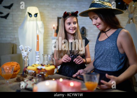 Portrait of two teenage girls wearing witch costumes setting up snack table at Halloween party in decorated room Stock Photo