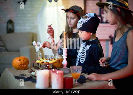 Portrait of three little kids wearing Halloween costumes standing at table with sweets during party in decorated room Stock Photo