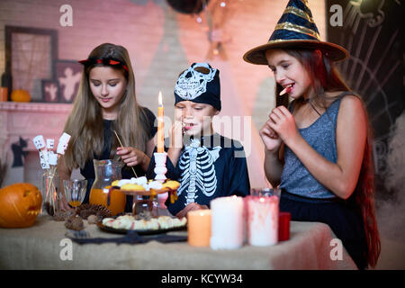 Portrait of three children wearing Halloween costumes eating sweets  and snacks during party in decorated room Stock Photo