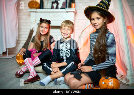 Portrait of three children wearing Halloween costumes posing looking at camera sitting on floor in decorated studio Stock Photo