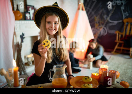 Portrait of pretty little girl dressed in Halloween costume posing with vampire cookie in decorated room, other children playing in background Stock Photo
