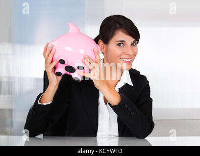 Young Happy Businesswoman Holding Pink Piggy Bank At Desk Stock Photo